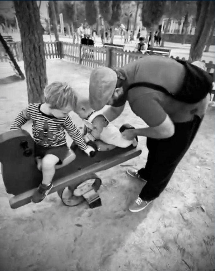 A father gently helping his child onto a wooden rocking horse in a park.